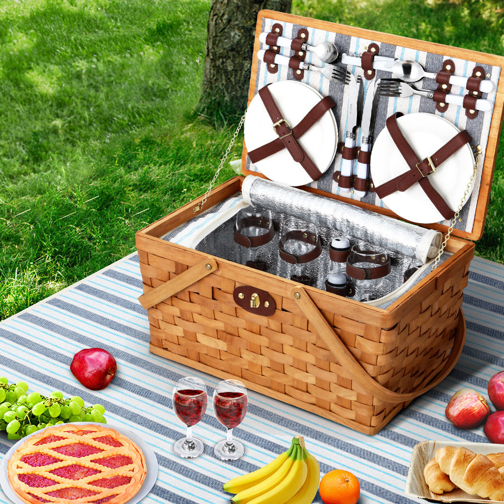 The Alfresco 4 Person Picnic Basket Set Wooden Willow with blanket and insulated bag, set on a picnic blanket with food and drinks around it, including a corkscrew, salt and pepper shakers, and stainless steel cutlery.